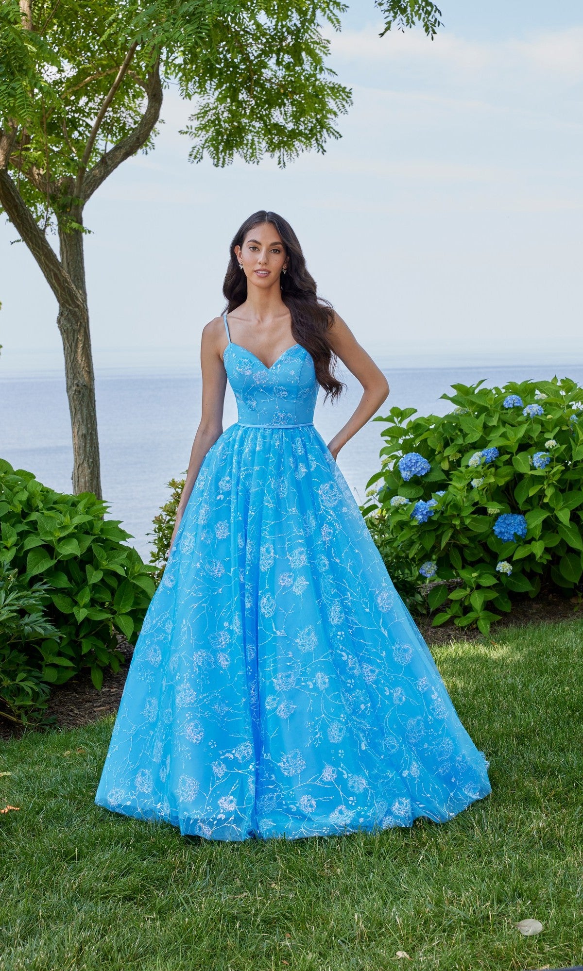 A woman in a French Blue Morilee 49153 long prom dress with an A-line silhouette and lace-up back stands on grass by green bushes and a tree, with water and sky in the background.